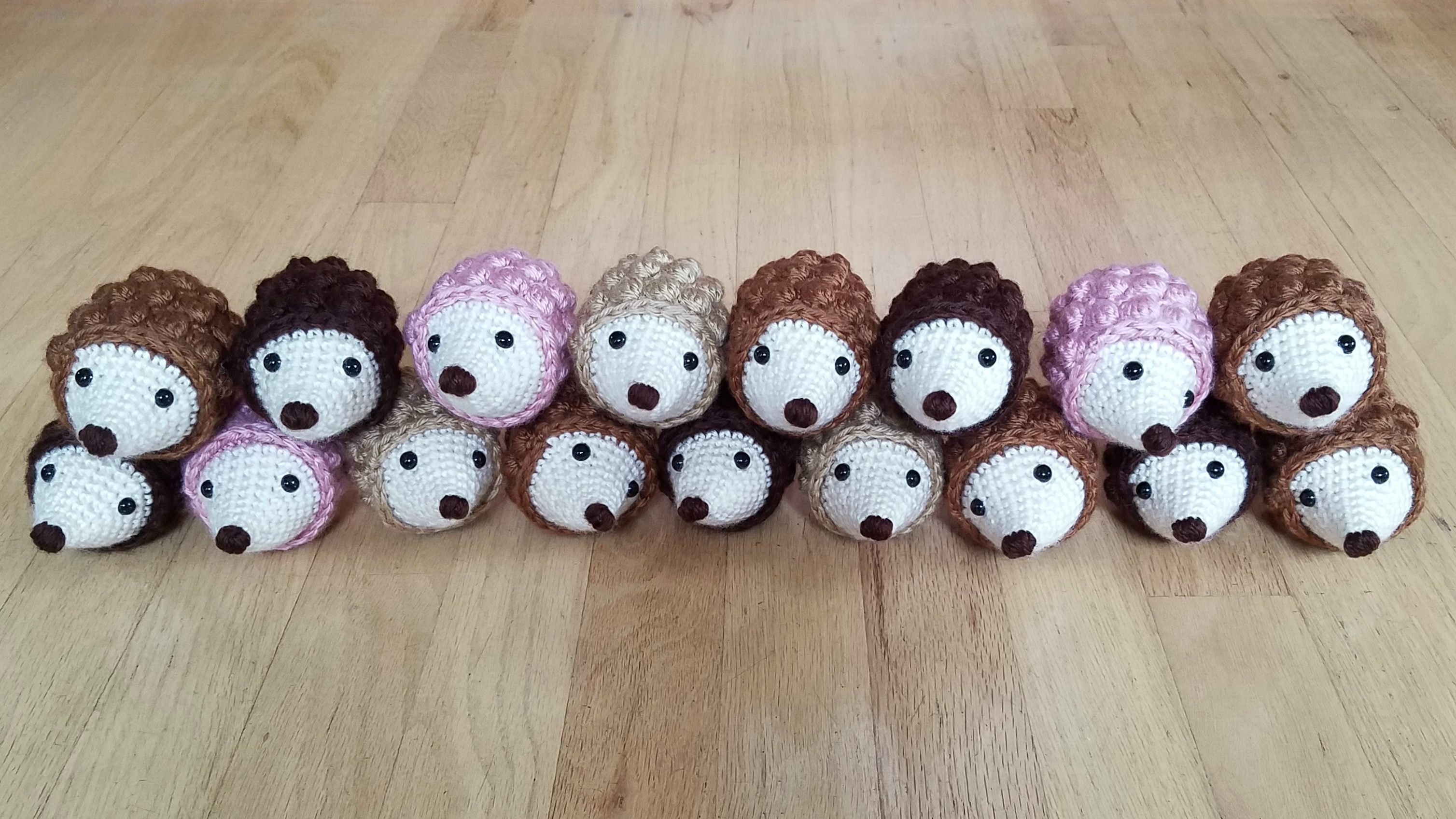Seventeen hedgehog amigurumi of various colors stacked on top of each other in two rows on a light-colored wood floor.