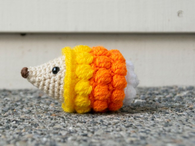 A left facing side view of a crochet hedgehog with a beige face and yellow, orange, and white bobble stitch spines.