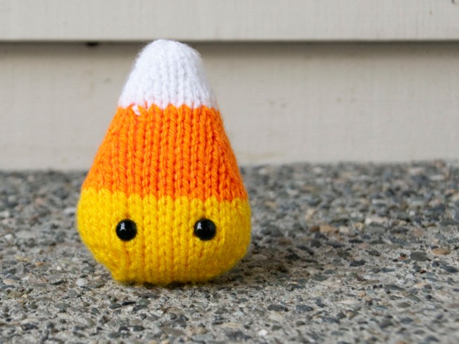 A knit candy corn with black eyes standing upright on pebble concrete.