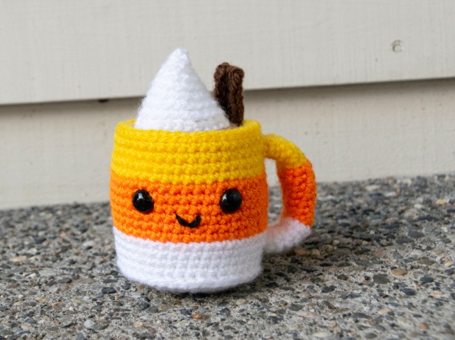 A crocheted cup of coffee with a yellow, orange and white striped cup, a white whipped cream dollop, and a crocheted cinnamon stick.