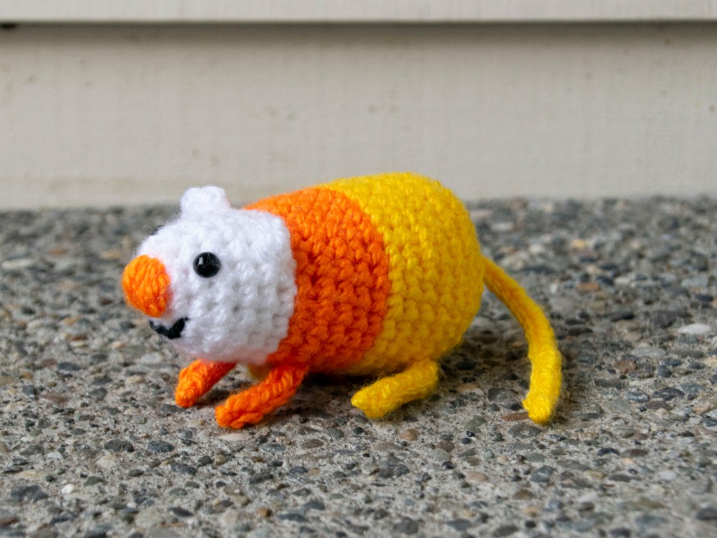 A crocheted candy corn rat with an orange nose, orange knitted front legs, and yellow knitted back legs and tail.